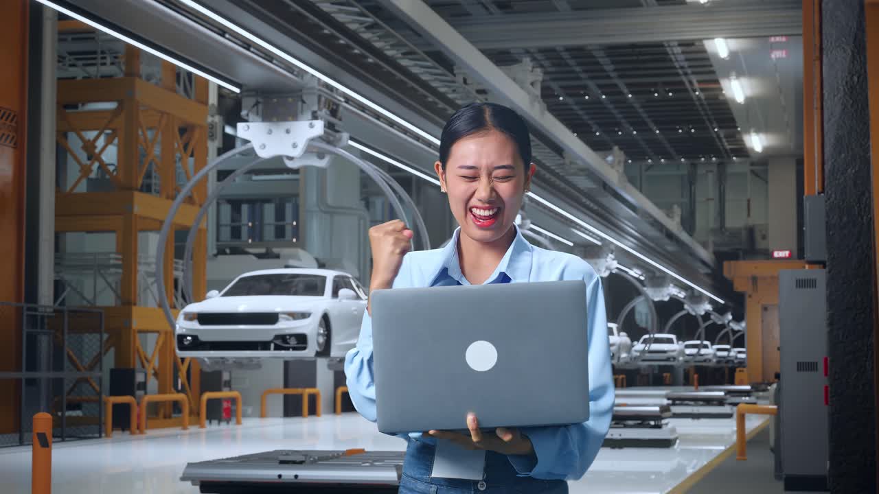 아시아 여성 직업 노동자가 ev 자동차 공장, 첨단 산업 시설에서 노트북을 들고 서서, 그녀는 소리치는 목표로 주먹을 들어 올립니다.