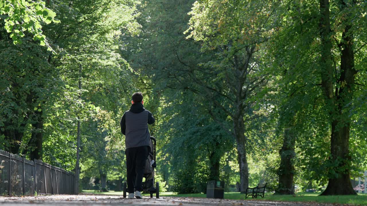 A young father walks with his baby in a pram through a tree-lined park on a sunny day, highlighting parenthood, family bonding, and peaceful outdoor lifestyle moments