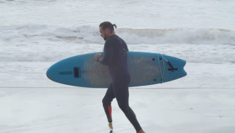 toma de seguimiento de un hombre discapacitado en traje de neopreno con pierna artificial corriendo a lo largo de la costa con tabla de surf