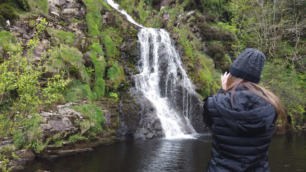 Young female photographer capturing scenic waterfall cascading through lush green irish landscape, documenting natural beauty of picturesque outdoor environment