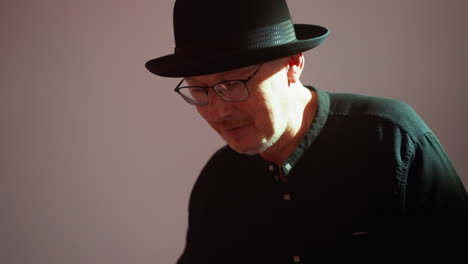 close-up of a man in a black outfit and hat against a soft pink background, looking down thoughtfully