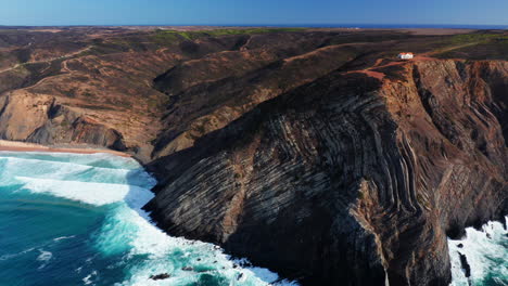 breathtaking aerial orbit shot of crashing waves against gigantic mountain cliffs with tiny house on mountaintop