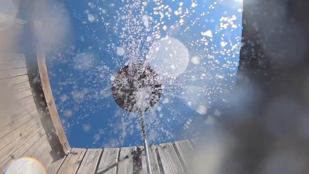 Gotas de agua cayendo de un cabezal de ducha elevado contra un cielo azul claro, enmarcado por paredes de madera desgastada. Experiencia refrescante al aire libre.