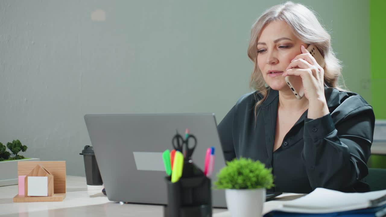 professional lady multitasks at modern office desk, managing phone call while working on laptop, surrounded by office tools, hot drink, and organized documents in productive workspace