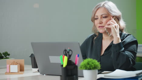 professional lady multitasks at modern office desk, managing phone call while working on laptop, surrounded by office tools, hot drink, and organized documents in productive workspace