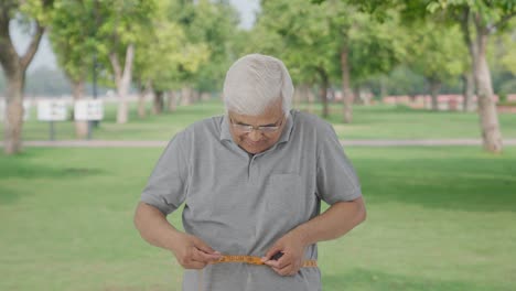 Feliz-Anciano-Indio-Comprobando-El-Tamaño-De-Su-Cintura-Usando-Cinta-Adhesiva-En-El-Parque