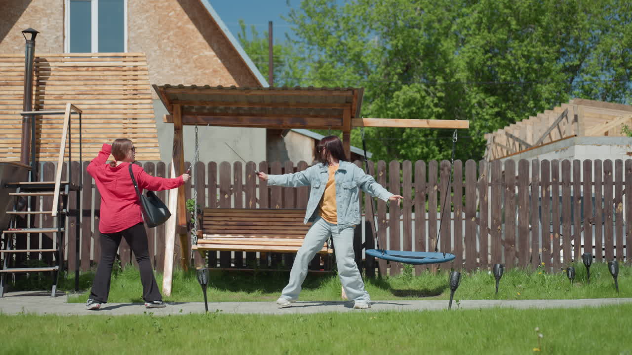 Two young women engage in playful fight beside wooden swing and suspended seat on grassy yard, one raising arm in jest, surrounded by fence, trees, lamps, and residential buildings