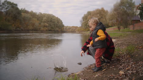 Frau-Und-Kleiner-Junge-Werfen-Steine-Ins-Wasser-Am-Ufer-Des-Flusses-Im-Wald.-Gemeinsame-Familienzeit-Am-Wochenende-Am-Herbsttag.-Glückliche-Mutter-Und-Kind