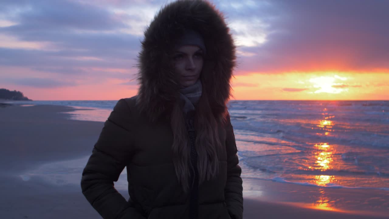 Young woman in winter clothes walks along the sandy shore of the Baltic sea beach at romantic sunset, medium shot pan left