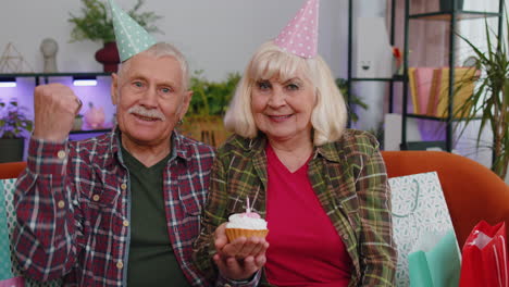 happy senior family man woman celebrating birthday anniversary hold cupcake makes wish at home