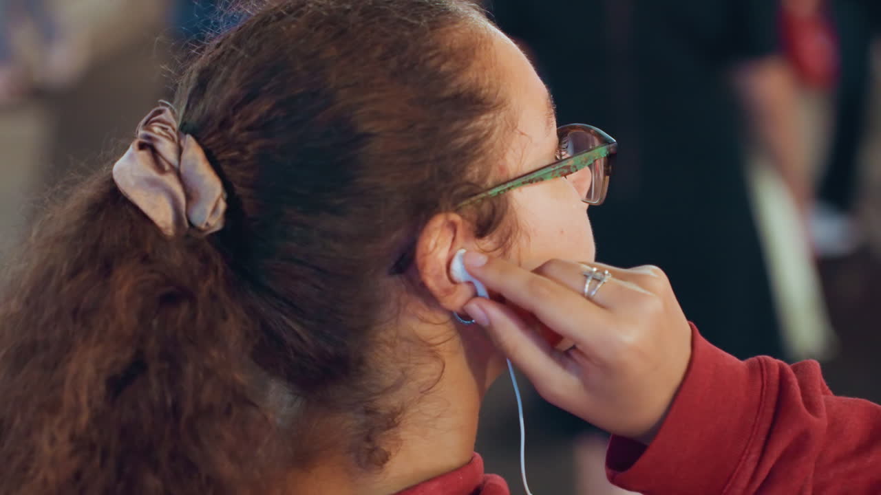 Un pendolare di colore con gli occhiali si sistema l'auricolare in un affollato attraversamento pedonale, tra luci al neon e una folla in rapido movimento, profilo concentrato con felpa con cappuccio ed elastico per capelli, frenesia urbana durante gli spostamenti notturni.