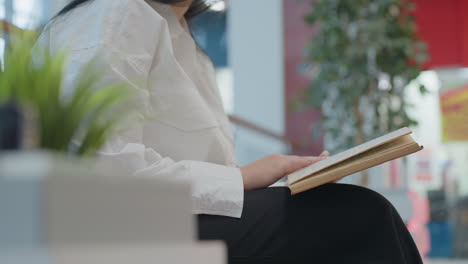 side view of woman dressed in white shirt and black pants flipping pages of book while seated, with blurred shopping mall background and soft lighting creating peaceful reading atmosphere