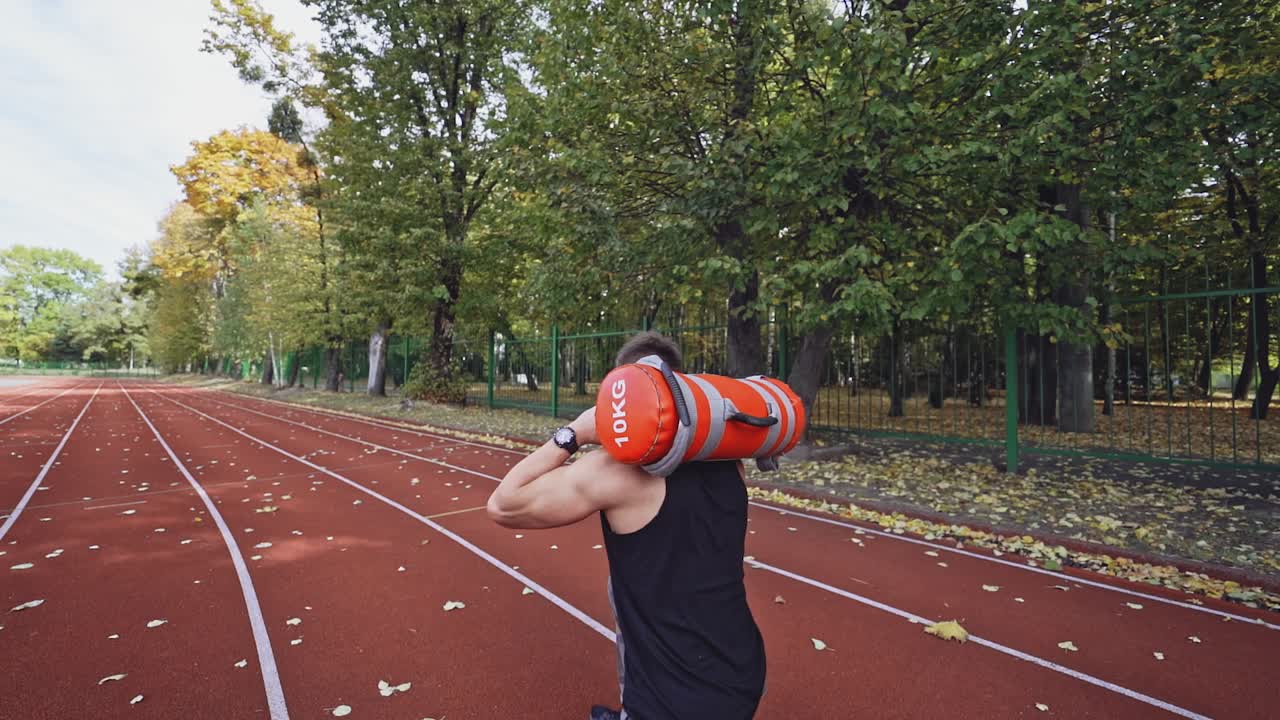 Strong athlete doing hard exercises on the open air. Sporty athlete training with weights on his shoulders on the stadium near the autumn park. Slow motion.