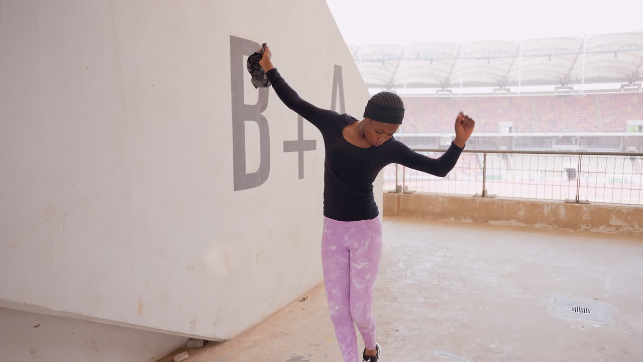 black woman vlogger dancing by stadium, wearing pink pants and black top while holding camera energetic movement along concrete corridor with graffiti BA, empty bleachers and overcast sky, candid