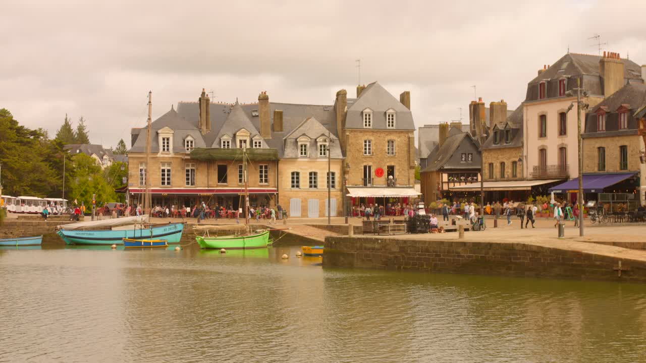 Rekaman menunjukkan Pelabuhan Saint-Goustan di Auray, Prancis dengan perahu-perahu berwarna-warni di air dan bangunan bersejarah yang berjejer di tepi pantai. Pencahayaan tengah hari menyoroti suasana yang dinamis dan aktivitas di area tersebut