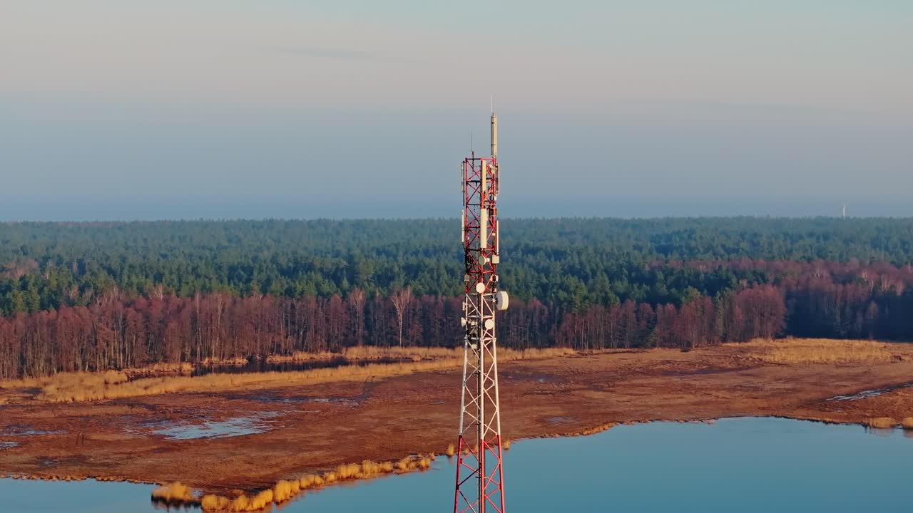 Drone che orbita fluidamente attorno a un'alta torre di comunicazione, alba primaverile sull'acqua