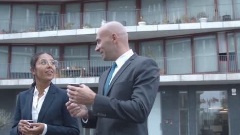 colegas de negocios sonrientes y confiados caminando cerca del edificio de oficinas, bebiendo café y hablando juntos