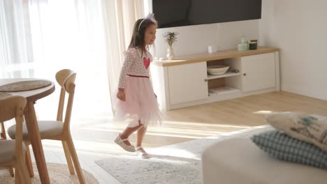 a young girl dances joyfully in a bright, cozy living room. she wears a pink dress with a heart design, moving playfully in the sunlit, minimalist space with neutral decor and soft natural light.