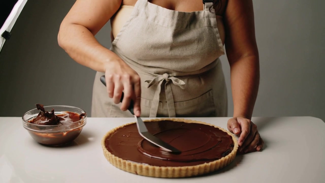Un fornaio con grembiule di lino immerge la spatola nella ciotola, spalma la ganache su una crostata, finendo il lavoro in studio