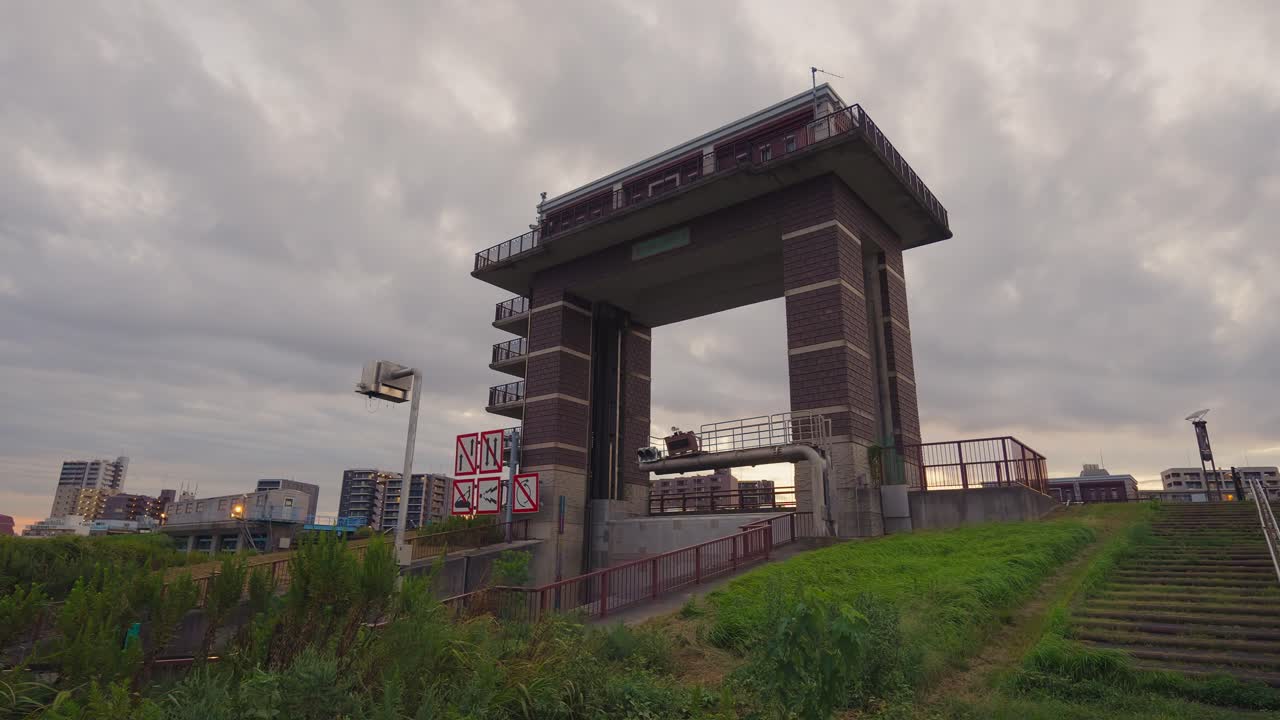 A low-angle shot of a large, industrial sluice gate structure against a cloudy dusk sky, with a grassy bank in the foreground