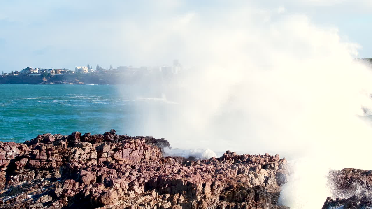 Crashing wave into jagged rocks of coastline sprays seawater into air, slomo