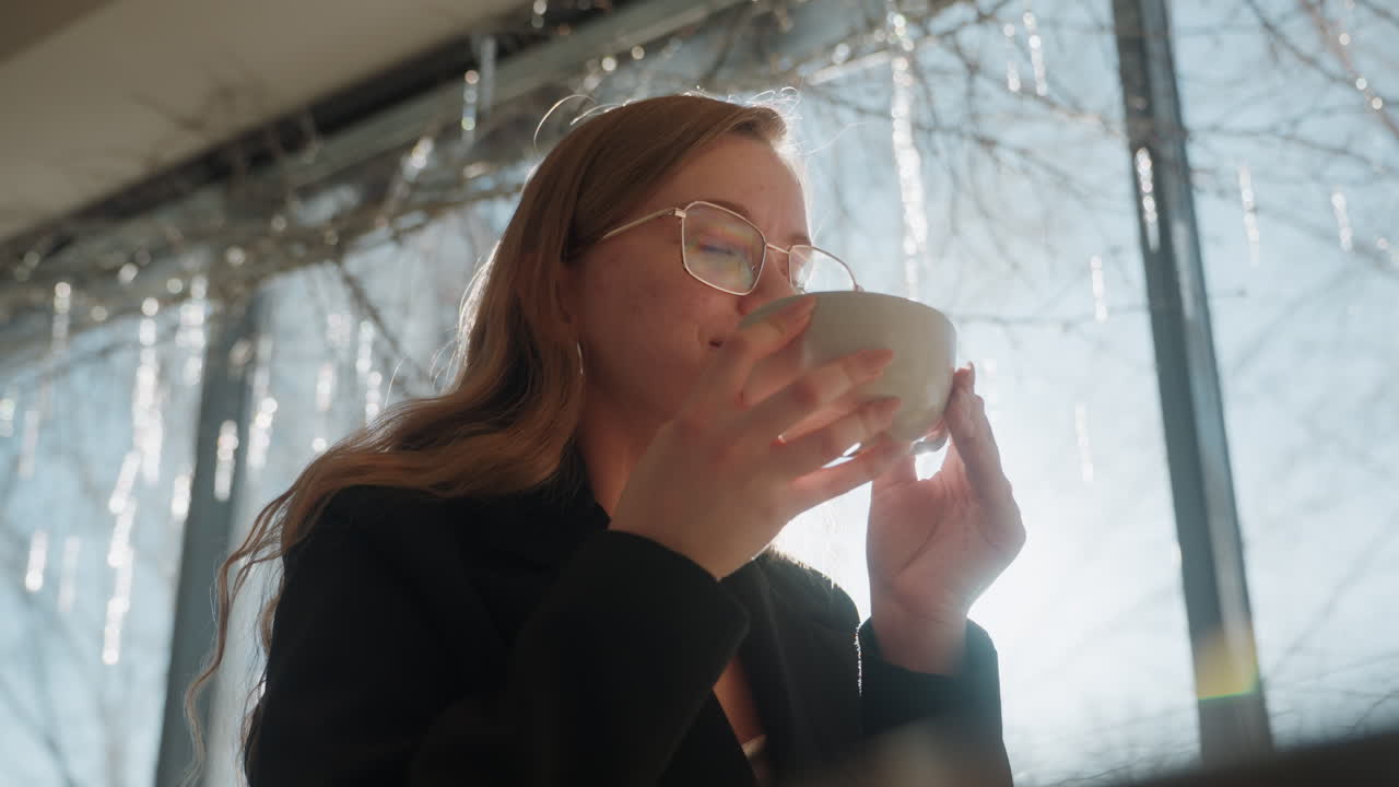 Hvid kvinde holder varm kop tæt på vinduet, modlys fra vintersolskin og istapper udenfor, mildt smil og tilfreds udtryk, porcelænskop gløder i morgenlyset, hyggelig festlig atmosfære.