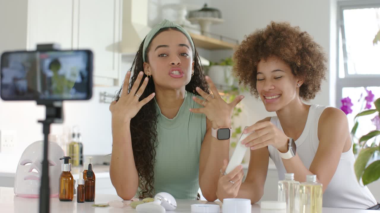 Two women filming skincare phone recording them, left touching face, right reading tube in kitchen