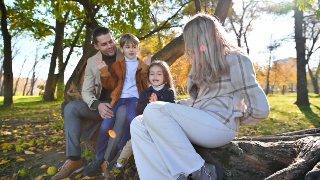Famiglia felice in un parco autunnale. Madre, padre, figlio e figlia seduti su un tronco d'albero, alberi ingialliti intorno. Movimento lento