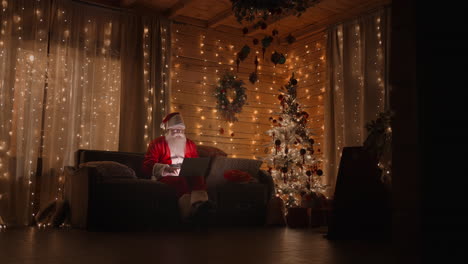 The-general-plan-of-Santa-Claus-at-home-is-sitting-on-the-couch-with-a-laptop-near-the-Christmas-tree-working-with-a-laptop