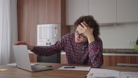 brooding serious curly freelancer man sit at table in comfortable home office room work on laptop looks concentrated thinking over business issue solution makes telecommute job