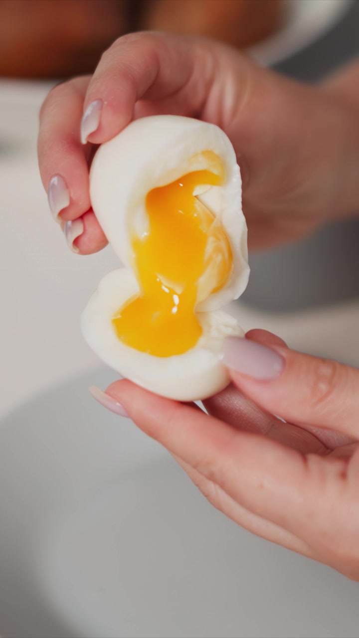 Woman hands gently hold and slightly rotate soft boiled egg revealing runny yellow yolk inside. close up view emphasizes texture of smooth white shell contrasted with glossy liquid center