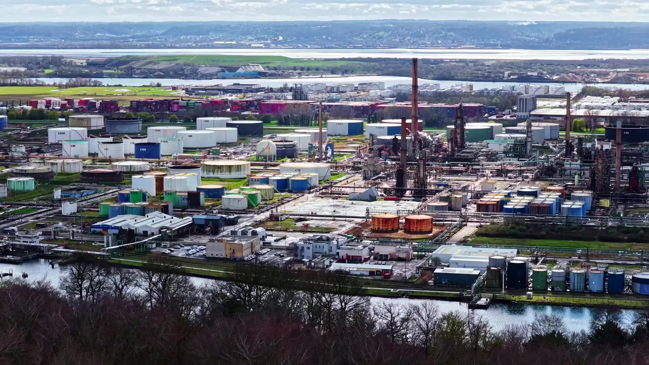 An industrial facility with numerous storage tanks and smokestacks stretches across the landscape, bordered by a river. Suitable for industrial or environmental projects.