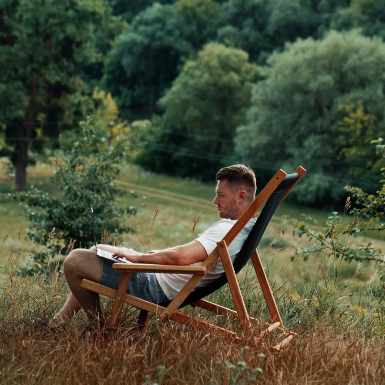 Nice place to work remote in the nature. Caucasian man works on his laptop sitting in a folding chair. Side view