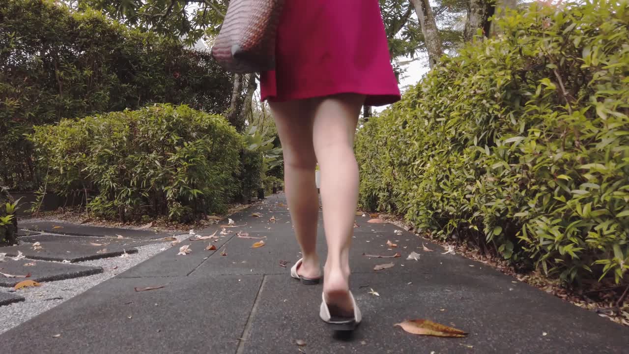 Una giovane donna asiatica in abito rosso con una borsa da spiaggia che cammina su un sentiero verso un paradiso balneare durante la vacanza estiva, al rallentatore.