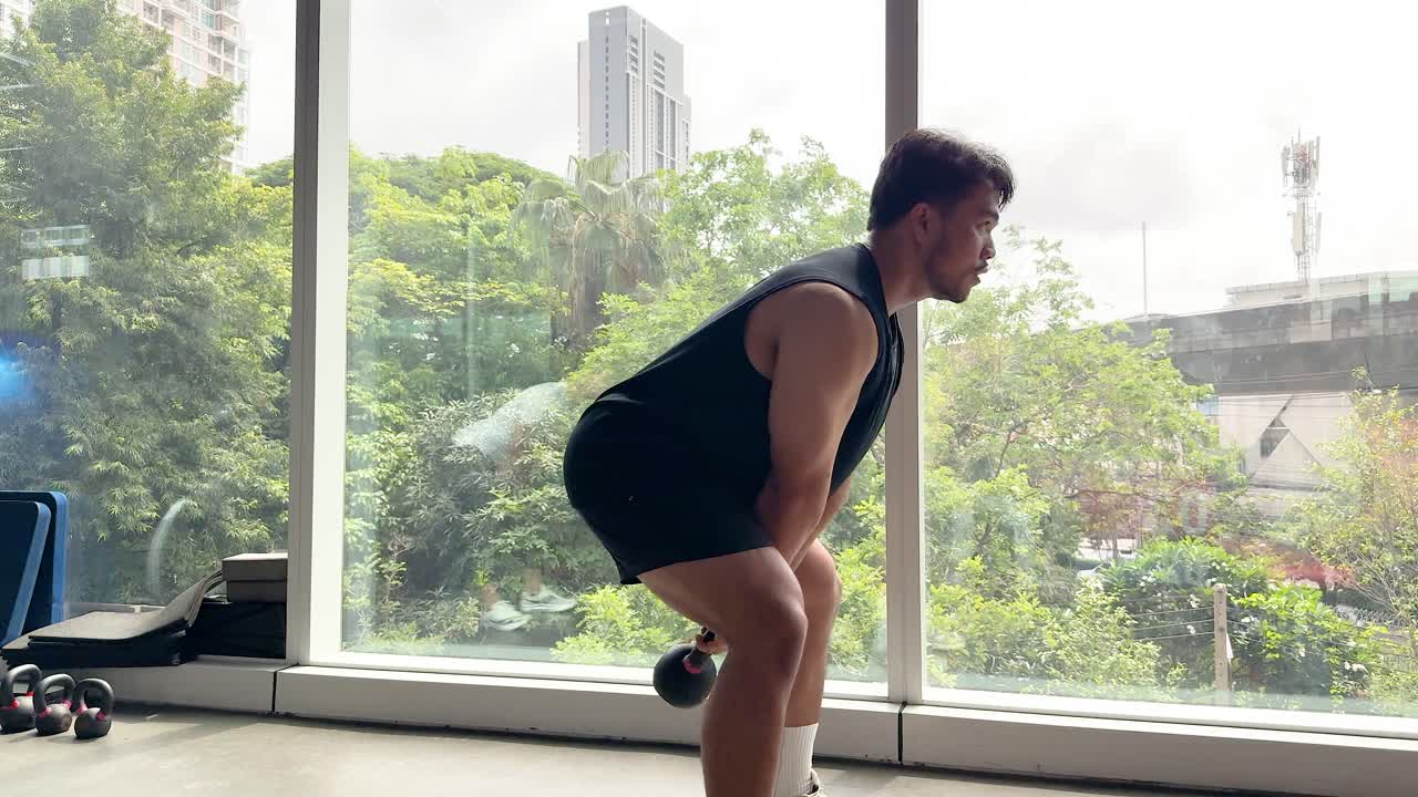 Two individuals perform kettlebell swings in a bright gym with large windows, emphasizing fitness and technique
