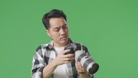 close up of asian photographer using a camera taking pictures and making stop hand sign while standing on green screen background in the studio