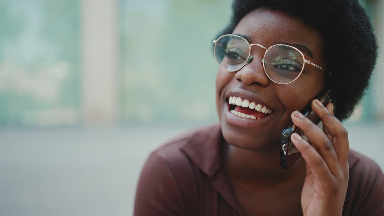 Primo piano di una ragazza africana con gli occhiali che sorride mentre parla al cellulare all'aperto