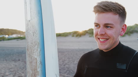 young man wearing wetsuit holding surfboard enjoying surfing vacation on beach as sun sets