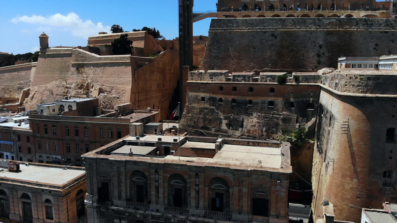 항구 옆 건물 꼭대기에서 흔들리는 몰타 국기의 공중 샷 - 위쪽 바라카 정원과 배경, 발레타, 몰타의 아름다운 하늘