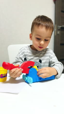 toddler kid sits at desk focused on his toy. adorable baby plays with plasticine and molding device. vertical video.