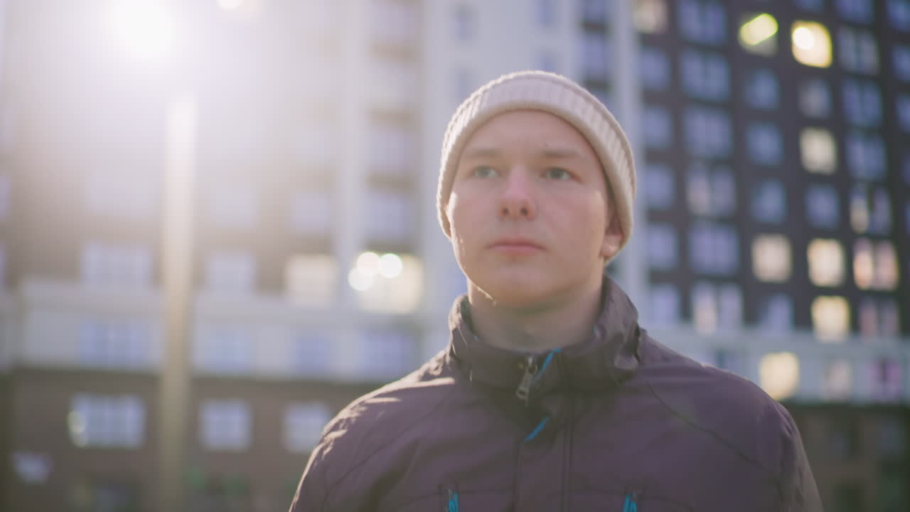 Retrato contemplativo de um homem branco ao entardecer usando gorro e jaqueta de inverno, retroiluminado por um poste de luz e janelas de apartamento, bokeh suave e reflexo de lente cinematográfico, olhar para cima, humor reflexivo, vizinhança