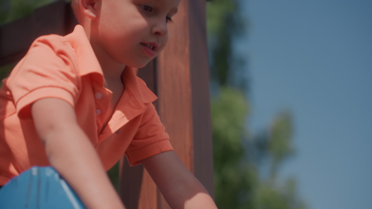 White Boy Walking Beside Metal Fence Orange Polo Turned Profile Thoughtful Expression Sunlit Yard Quiet Pause Subtle Gestures Suburban Background Cinematic Color Grading Introspective Summer Scene