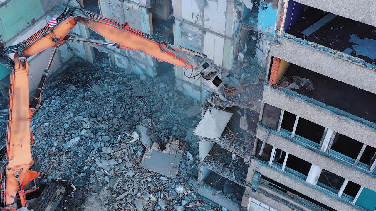 Excavator crashing floors of an old building and dusty debris falling down. Disassembling of collapsed structures. View from above.