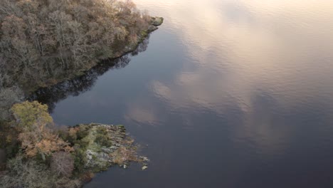 Imágenes-Aéreas-De-Drones-De-Un-Amanecer-De-Otoño-Con-Nubes-Reflejadas-En-Loch-Lomond-Con-Un-Bote,-Un-Pájaro-Volador-Y-Un-Bosque-De-Hoja-Ancha-Abrazando-Sus-Costas-En-Loch-Lomond-Y-El-Parque-Nacional-Trossachs