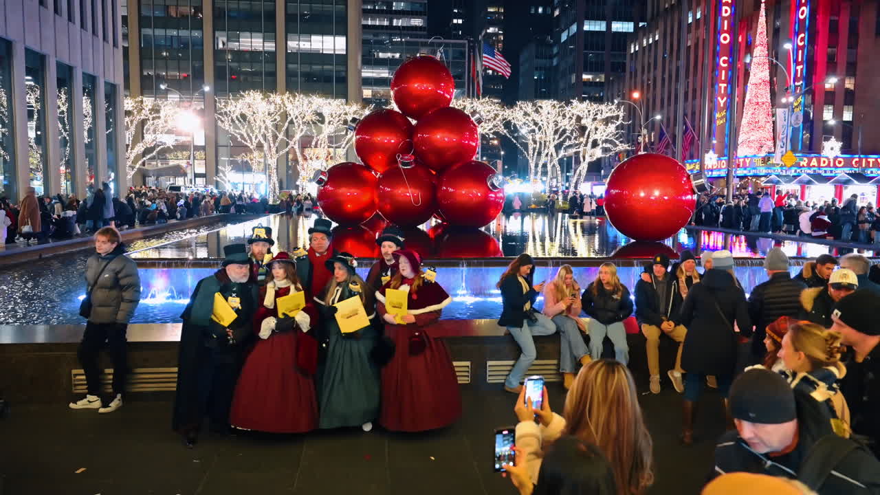New York, USA, 5 December 2025: Victorian Carolers in City Square Surrounded by Spectators. The carolers in full Victorian dress stand by a fountain and red ornaments, observed by onlookers taking photos in the evening