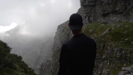 a man shown from behind standing on a mountain and looking out over the misty ridge of the mountains in front of him