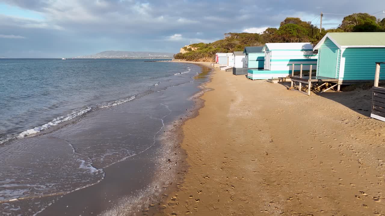 Steady camera moves past vibrant beach huts on sandy shoreline, morning sunlight, gentle sea waves