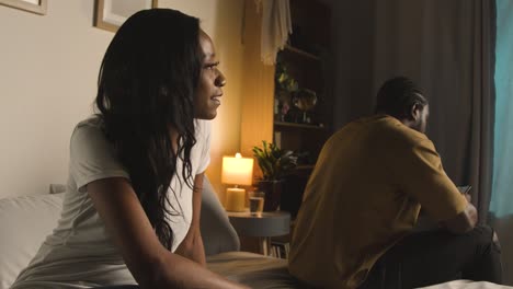 young couple at home at night in bedroom with woman talking to man scrolling internet on mobile phone