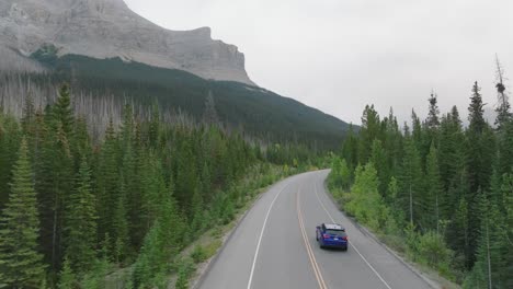 Carretilla-Aérea-En-La-Conducción-De-Automóviles-En-La-Carretera-Entre-El-Bosque-De-Pinos,-Las-Montañas-Rocosas-Canadienses-En-El-Fondo,-El-Parque-Nacional-De-Banff,-Alberta,-Canadá