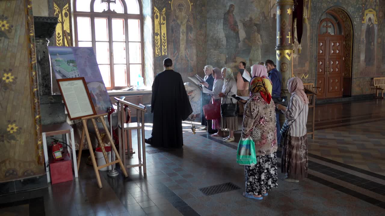 Ukraine, Kyiv, Pechersk Lavra, interior of the famous church, a young priest in the front and some people, primarily women, praying behind him. a day time ceremony in the cosy historical church.